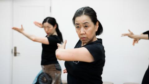 Women practise flamenco dancing. They are wearing black T-shirts and have their hair tied back in ponytails. 