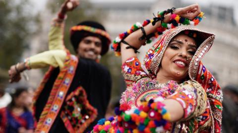 A man dancing on the left in black traditional Indian clothing with a woman on the right with a colourful Indian outfit on also dancing. 