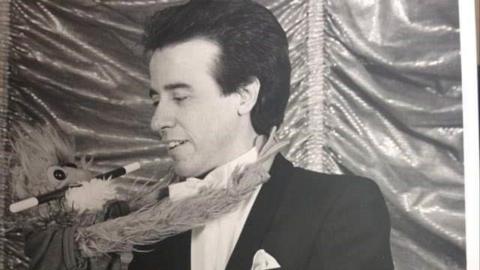 A black and white image of Reg Martin as a younger man performing magic. He is holding a plush doll with a magic wand in its mouth. Reg is looking at the doll. 