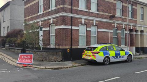 A police car and a road closed sign next to a cordon on a residential street in Plymouth.