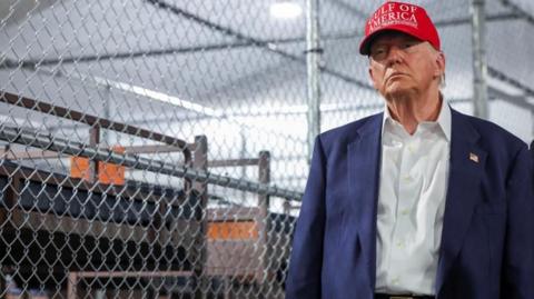 Trump stands in blue suit, red cap next to chain link fence and rows of bunk beds