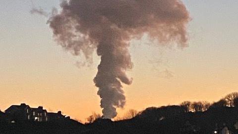 A large cloud of white steam billows into the orange evening sky, with houses and a tree-lined hill silhouetted on the horizon. 