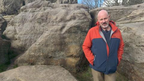 Roger Barton, treasurer for FTWRC, pictured in the Tunbridge Wells and Rusthall Commons. Roger is wearing a grey jumper and red/navy coat. He is bald, and is standing in front of natural sandstone formations.