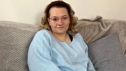 A woman wearing a light blue jumper with glasses and brown shoulder-length hair sitting on a sofa.