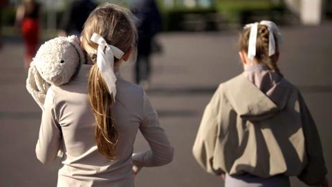 Two young girls walk alongside each other with their backs to the camera. They're wearing grey with their hair tied back in a bow. The one on the left is carrying a large stuffed toy rabbit.