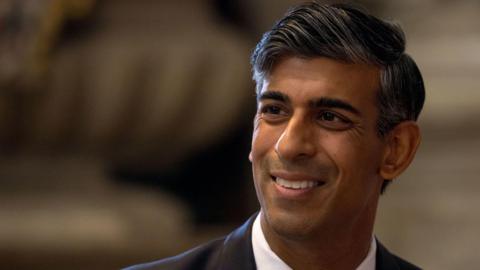 A headshot of former prime Minister Rishi Sunak. He is smiling and looking away from the camera.