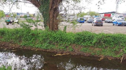 A shallow brook runs adjacent to a small car park with Hereford FC's stadium in the background.