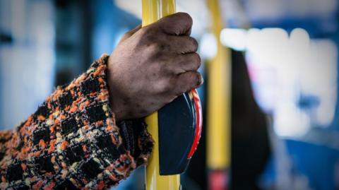 The hand of a woman wearing a tartan overcoat grips the bar of a bus just above the stop bell push.