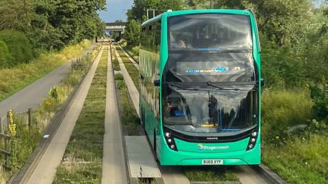 A double-decker bus on a guided busway heading towards the viewer along concrete tracks and kerbs in 2021. It has bright green livery. It is branded Stagecoach. It is travelling along a concrete track. On its right is a cycle and walkway. On either side are trees. 