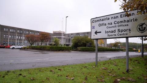 A sign pointing to Comhairle nan Eilean Siar's offices and a sports centre. A large grey municipal building is in the background.