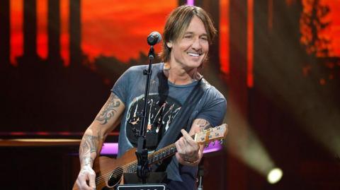 Keith Urban is playing on stage. He has brown hair down past his ears and is wearing a grey/blue T-shirt and playing an acoustic guitar and smiling. Behind him the red/black backdrop of the stage can be seen, in front of him is a microphone on a stand.