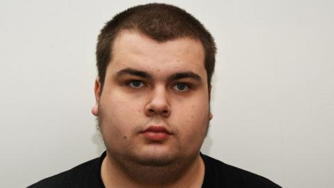 Head and shoulders police photo of Cameron Finnigan, photographed against a featureless white wall. He has short hair and is wearing a black t-shirt.