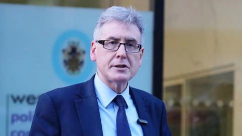 A man in a navy blue suit and blue tie with black rimmed glasses
