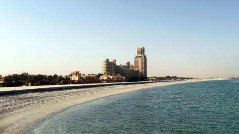 Ras Al Khaimah beach, which is sandy and has a clear blue sea. There are a few tall buildings scattered along the coast.