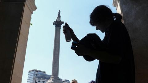 A person stands silhouetted in the shade at the side of Trafalgar Square, and holds misting spray up to their head to help cool themselves down, with Nelson's column in the background and people and a bus walking past.