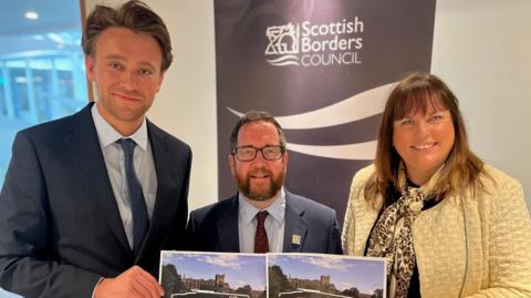 Two men and woman looking into the camera. They are standing in front of a Scottish Borders Council poster. They are smartly dressed - the men in suit and tie, the woman with a yellow jacket and neck scarf.  