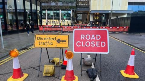 A road closure sign with a number of orange cones around it. 