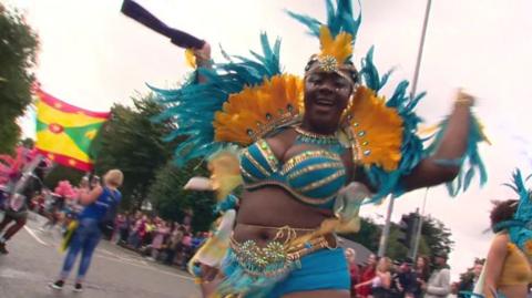 A woman in a vibrant blue and orange costume with matching feathers in the parade and crowds gathered to watch in the background
