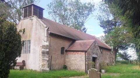 The churchyard at Yapham. The main church building is in the centre of the image with a large church tower in the background. A graveyard is visible in the foreground with large shrubs and iron railings surrounding the site.
