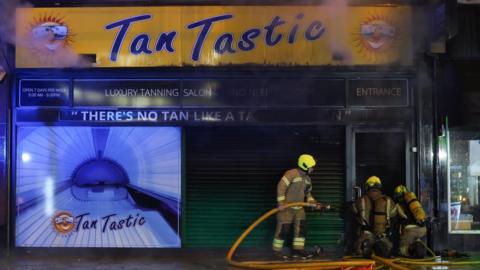 Three firefighters stand with a hose at the entrance of a shop front with a large yellow sign reading "Tan Tastic" in blue.