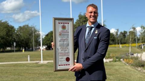 Dan Burn standing outside holding the framed scroll. He is wearing a blue suit and tie and has a small, gold medal attached to his blazer. He's smiling into the camera.