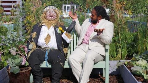 A man in a beige suit, Hamza Yassin, sits on a garden bench facing a scarecrow. 