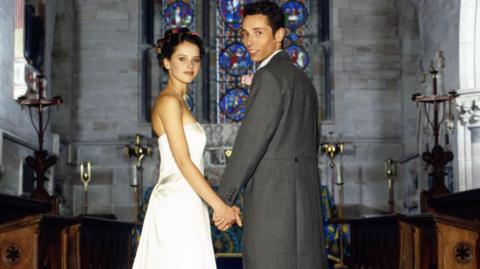 A bride and groom stand hand in hand inside a church, a large stained-glass window behind them. The bride has dark hair piled up and is wearing a strapless white dress, standing side on, looking to camera. The groom has his back to the camera and is turning around and smiling. He is wearing grey tails and trousers. Wooden pews and brass church ornaments can be seen around them.