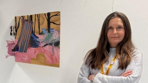 Belinda Downing is standing next to her artwork. She is wearing a grey jumper and is looking directly at the camera with her arms crossed.