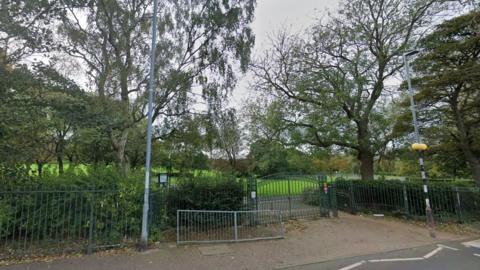 One of the entrances to Saltwell Park in Gateshead. A zebra crossing (just in shot on the right hand side) leads to a green pair of gates set into a green metal spiked fence. Behind it is an expanse of green grass and several trees. There is a buckled, grey metal street barrier just outside the gates and a wooden sign to the left. It is an overcast day but the trees are in full leaf.