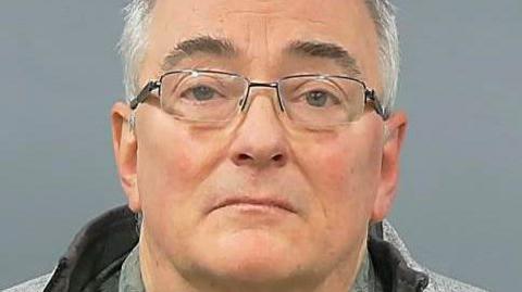Gerard Hutchings faces the camera in a police custody photo. He has short, grey hair and wears rectangular spectacles and a grey fleece.