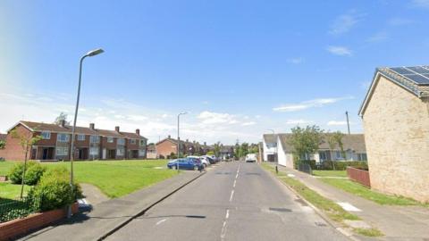 Tithe Barn Road. There are rows of two-storey houses and parked cars on each side of the two-lane road. 