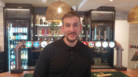 A man with brown hair and a beard stood in front of lager taps and a bar. 
