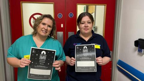 Two women are standing in a corridor in front of red double doors and each is holding black and white flyers advertising "The Great Tommy Sleep Out". One of the women has short blond hair and is wearing a turquoise medical top and the other has brown hair and is wearing a dark blue medical top.