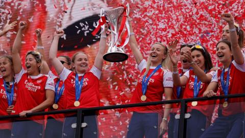 Arsenal's Kim Little and Leah Williamson and teammates celebrate with the trophy during the victory celebrations by lifting the cup. Red and white confetti is raining down.