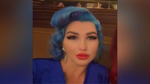 Tania Williams is inside looking to the camera. She has wavy blue hair and make-up on, including bright red lipstick and black mascara. She is wearing a dark blue dress.