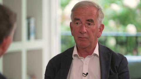 Robert Harris sits in a bright room with a balcony and trees visible in the background. He wears a dark blazer and a white shirt.