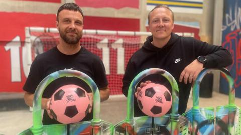 Two men holding pink footballs stand in front of a football net