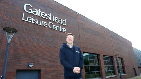 Robert Waugh standing outside Gateshead Leisure Centre. He is smiling and wearing a blue hoodie. Gateshead Leisure is behind him. It is made from brown brick and there are three windows to waugh's right.