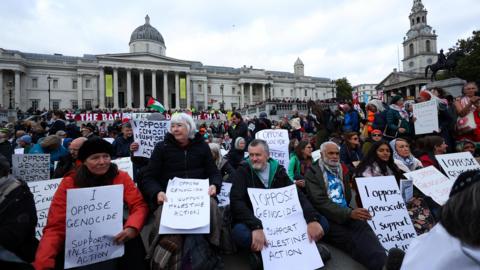 Protesters holding signs saying "I oppose genocide, I support Palestine Action" sit in front of the National Gallery in central London. 