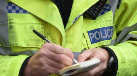 A close-up image of a police officer wearing a high-vis police jacket while writing in a small notebook. Only the officer's hands and torso can be seen.