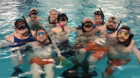 A mixed gender group of people pose with their arms around each other in a swimming pool. Everybody wears black snorkels, swimming caps and masks. The three people at the front can be seen wearing bright orange swimming trunks and flippers.