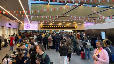 A train station concourse filled with a large crowd of people, many carrying luggage or backpacks. There are digital screens on the walls showing information.