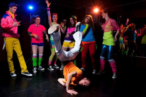 A group of young people dressed in 80s gear dancing with a young man breakdancing in the front