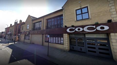 An image of a former nightclub.  It is a cream brick coloured building which fronts the pavement.  It has brown painted windows and the name CoCo in white.