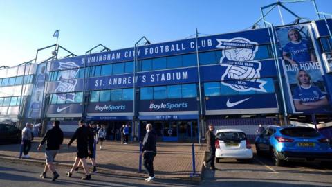 The front of a football stadium. The entrance to St Andrew's Park, the Birmingham City ground. The building is blue with large windows and a sign reading "Birmingham City Football Club". People are walking on the pavement near the entrance and cars are parked on the right.