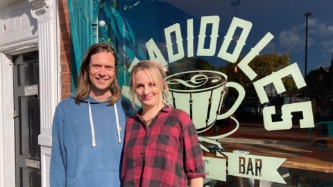 A man in a blue hoodie is standing next to a woman in a red and black oversized shirt. both are standing outside a large window that says Paradiddles music cafe bar 