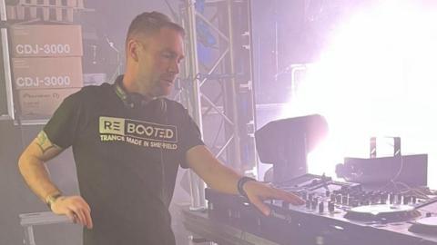 DJ Danny Stubbs wearing a black T-shirt, stood at a dj desk. He appears to be stood on a stage, with bright lights shining from the area to the front.