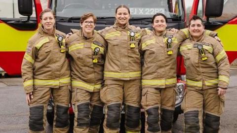 Five women in fire uniforms stand arm-in-arm