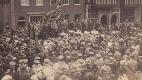 A black and white image of a two-minute silence in Farnham on 1 May 1916 with crowds of people and dignitaries with flags on stage in front of a big building