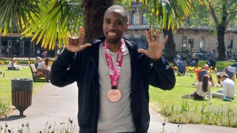 A man wearing a black hoodie, a beige sports top and a medal with a pink ribbon stands in front of a palm tree, in a park. He holds up seven fingers and smiles.